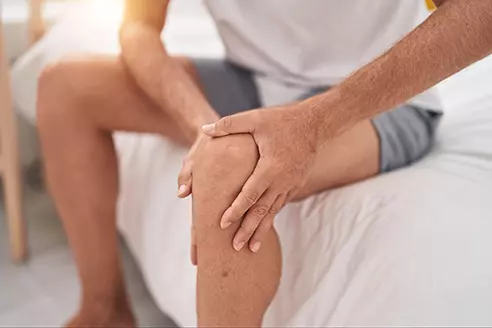 Man in grey shorts sitting on a bed holding his knee in pain, a common condition treated with PRP therapy.