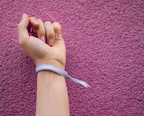 Raised fist wearing a transgender pride wristband against a pink textured wall, representing gender identity and transgender care.